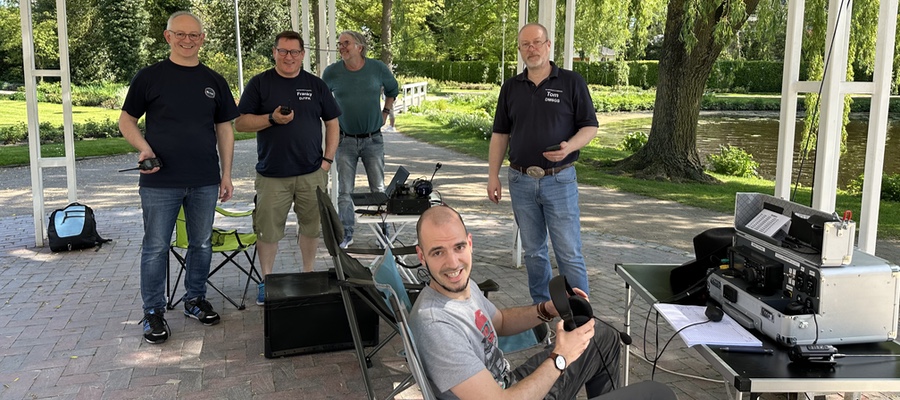 Wir blicken in einen Pavillon in einem Park. Vier Personen stehen im Hintergrund, eine sitzt im Vordergrund. Auf einem Camping-Tisch steht ein Funkgerät in einem Koffer.