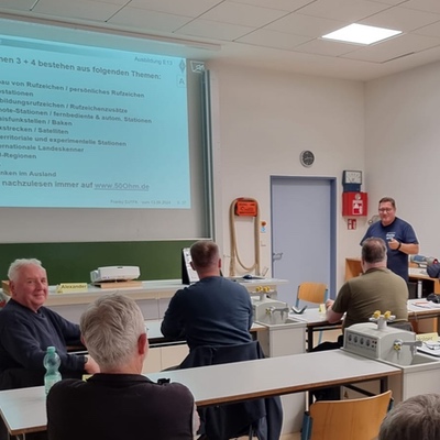 Franky steht vorne in einem Klassenraum und erklärt. Der Beamer zeigt eine Folie mit viel Text. Im Vordergrund sitzen Teilnehmende an Tischen.