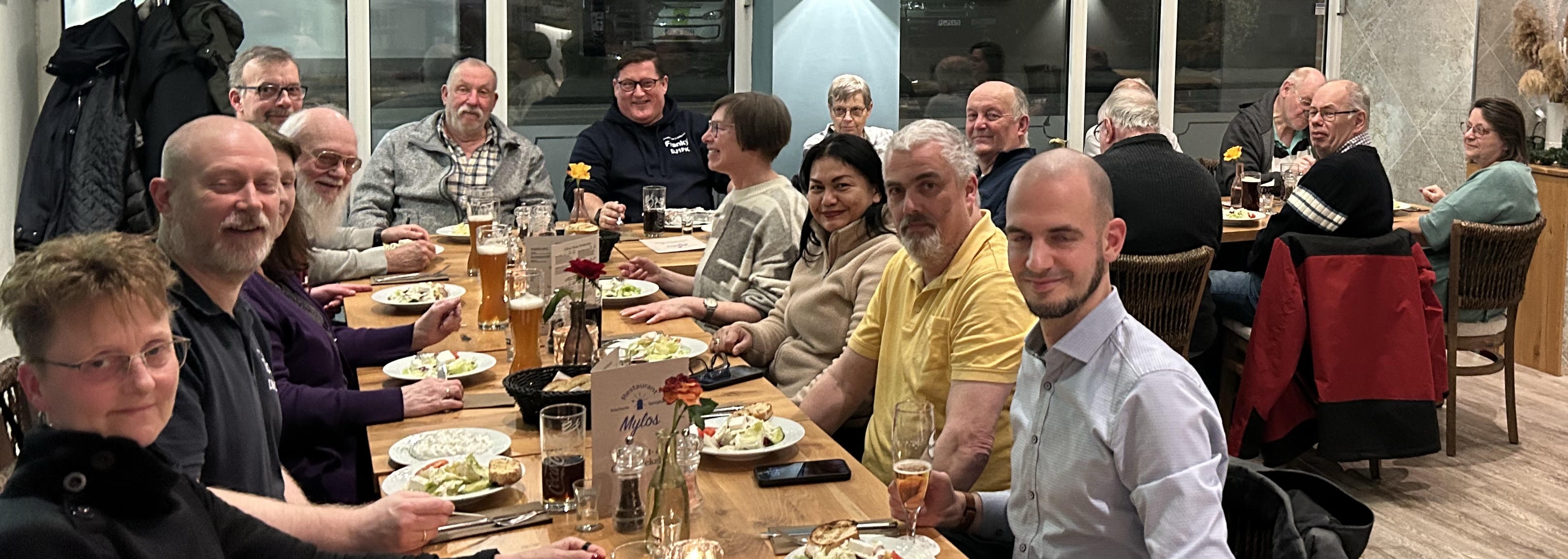 Blick über einen langen Tisch in einem Resteaurant, an dem viele Menschen sitzen und in die Kamera lächeln