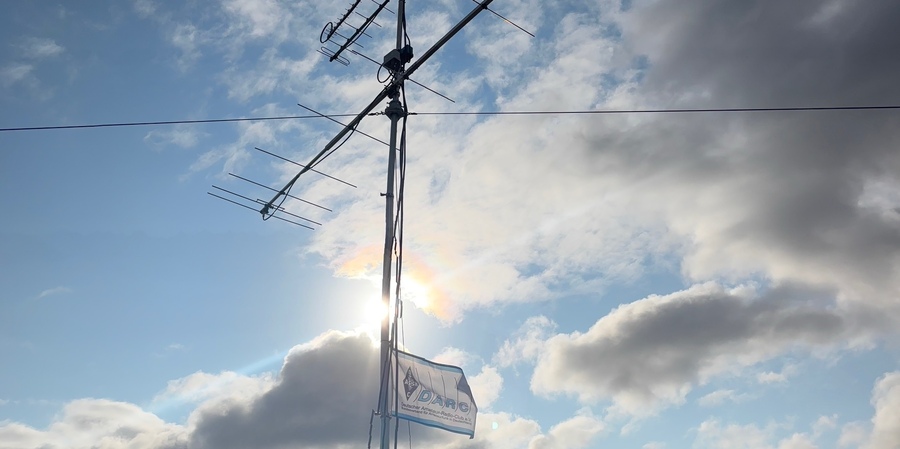 Amateurfunkantennen an einem Mast vor blauem Himmel; darunter weht eine DARC-Flagge.