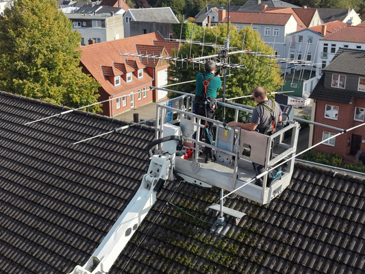 Drohnenaufnahme von zwei Personen im Korb des Hubsteigers, die auf dem Dach des Stadtwerkehauses am Antennenmast der Klubstation arbeiten.